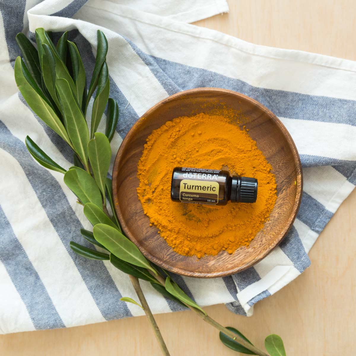 Bottle of doTERRA Turmeric oil surrounded by dry turmeric powder, a wooden dish, green leaves, and a blue striped dish towel. Does Turmeric essential oil have health benefits. When taken internally, Turmeric oil can support the nerv